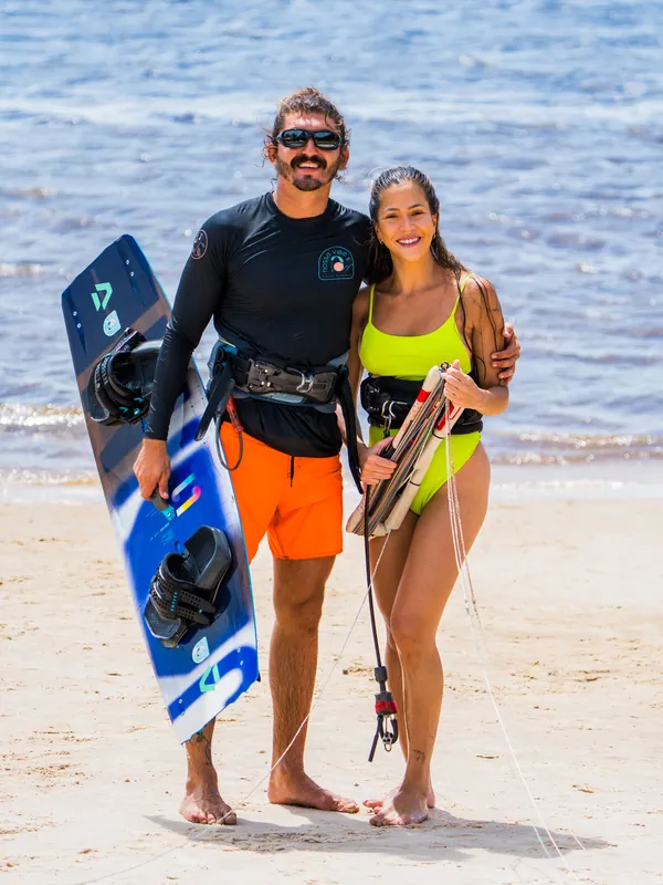 Um homem com a camisa da Nossa Vibe e a prancha, abraçado a uma mulher que segura a barra do kite, na beira da praia