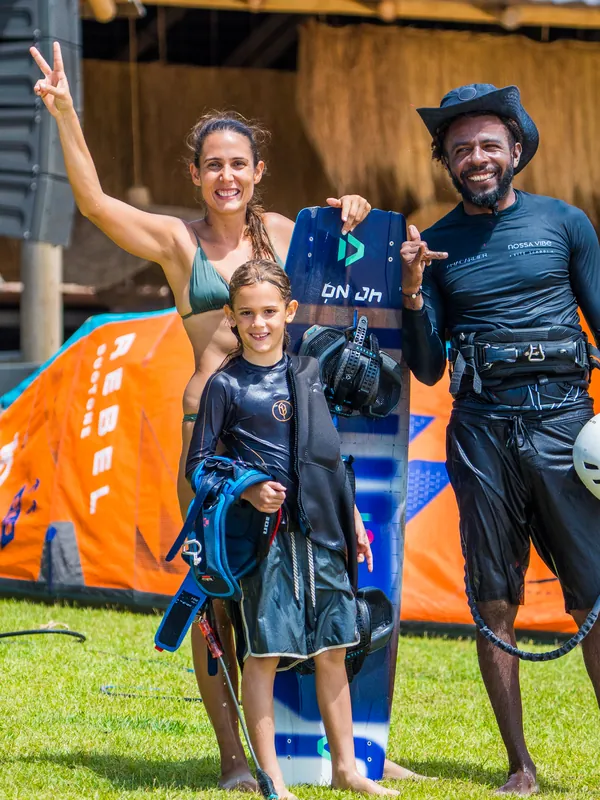 Uma mulher, uma menina e um homem com a camisa da Nossa Vibe sorrindo no gramado da escola, com a prancha e o kite laranja ao fundo