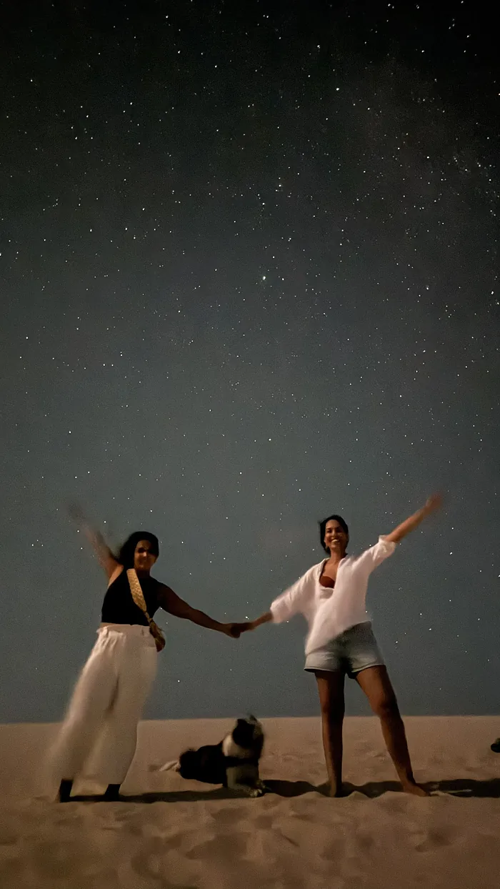 Duas amigas de braços abertos numa duna, sob o céu estrelado